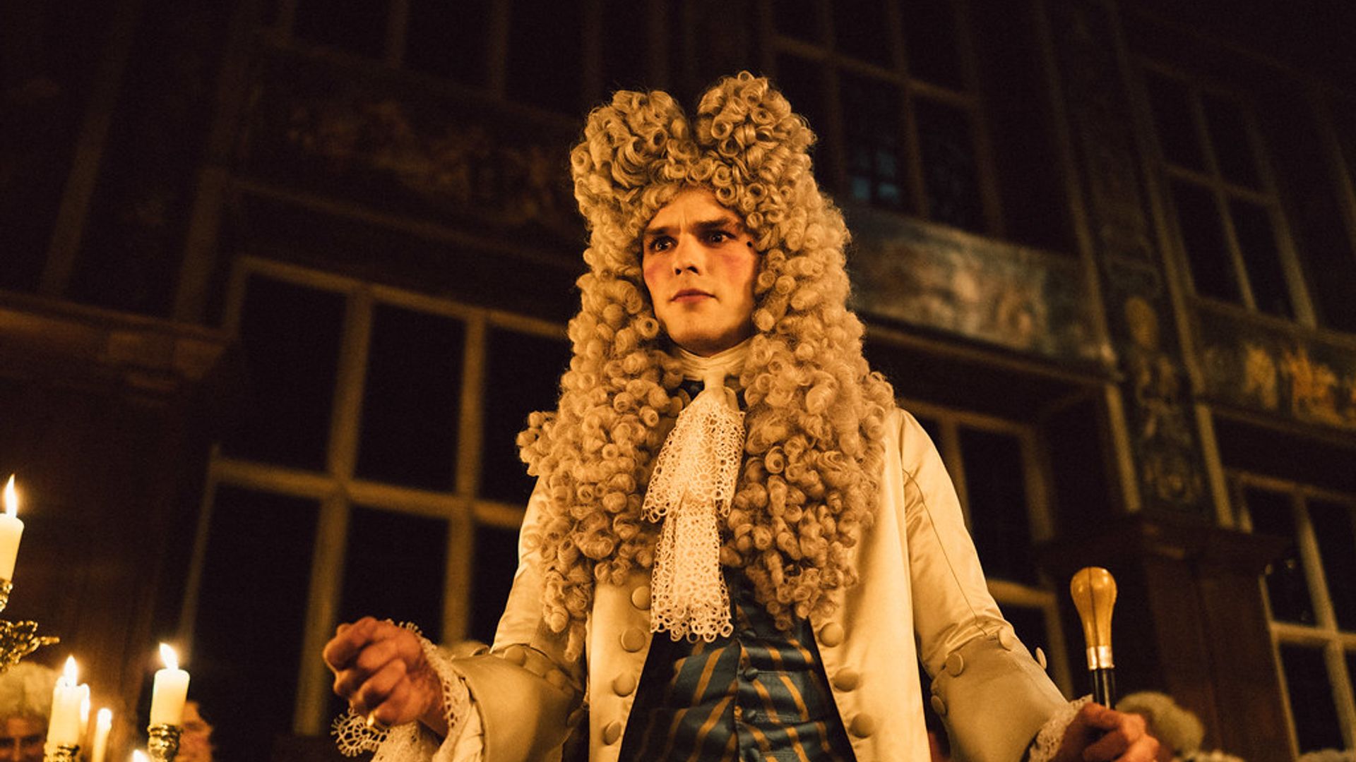 Nicholas Hoult in a wig and 18th-century period costume with a walking stick stands in a candlelit hall