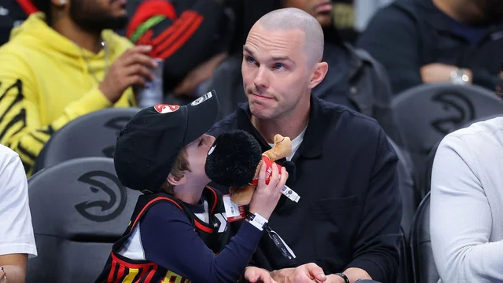 Nicholas Hoult sits in the stands at a basketball game next to his son who is holding a toy