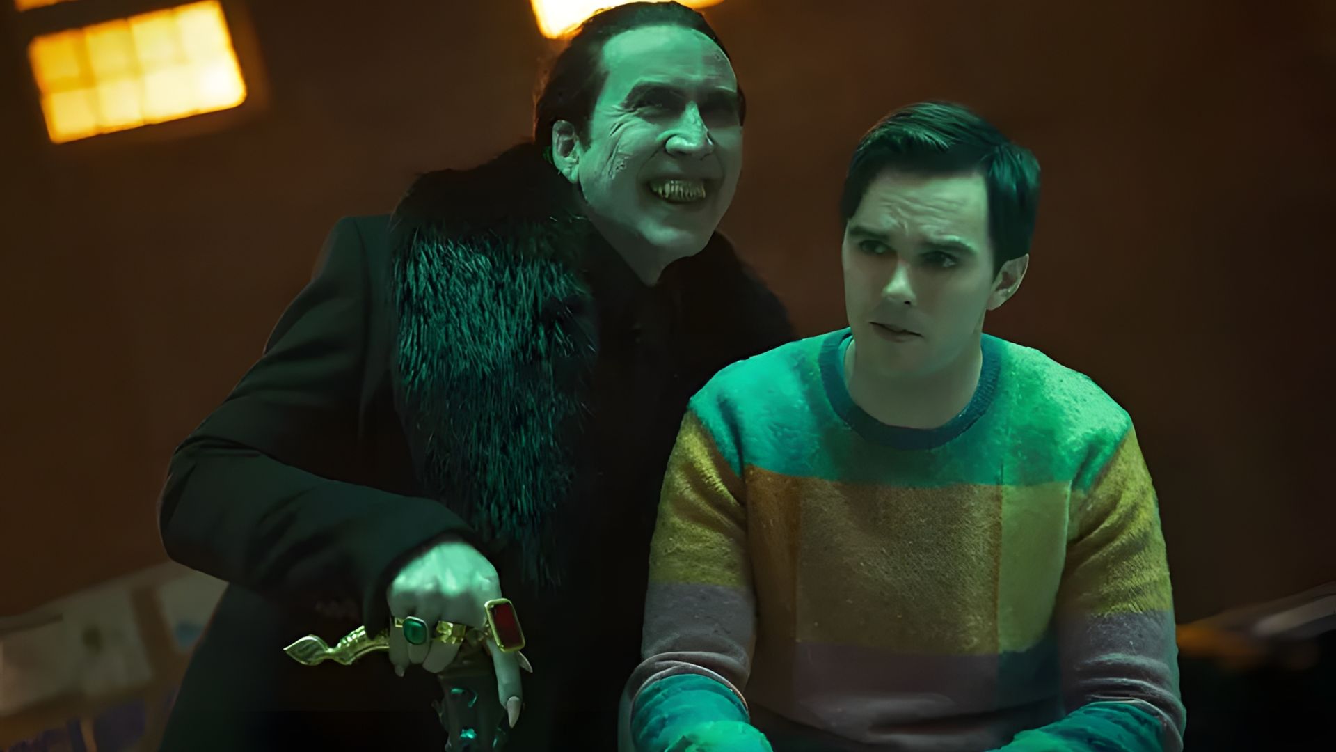 Nicolas Cage in black clothing with rings menacingly looms over Nicholas Hoult in a colorful sweater