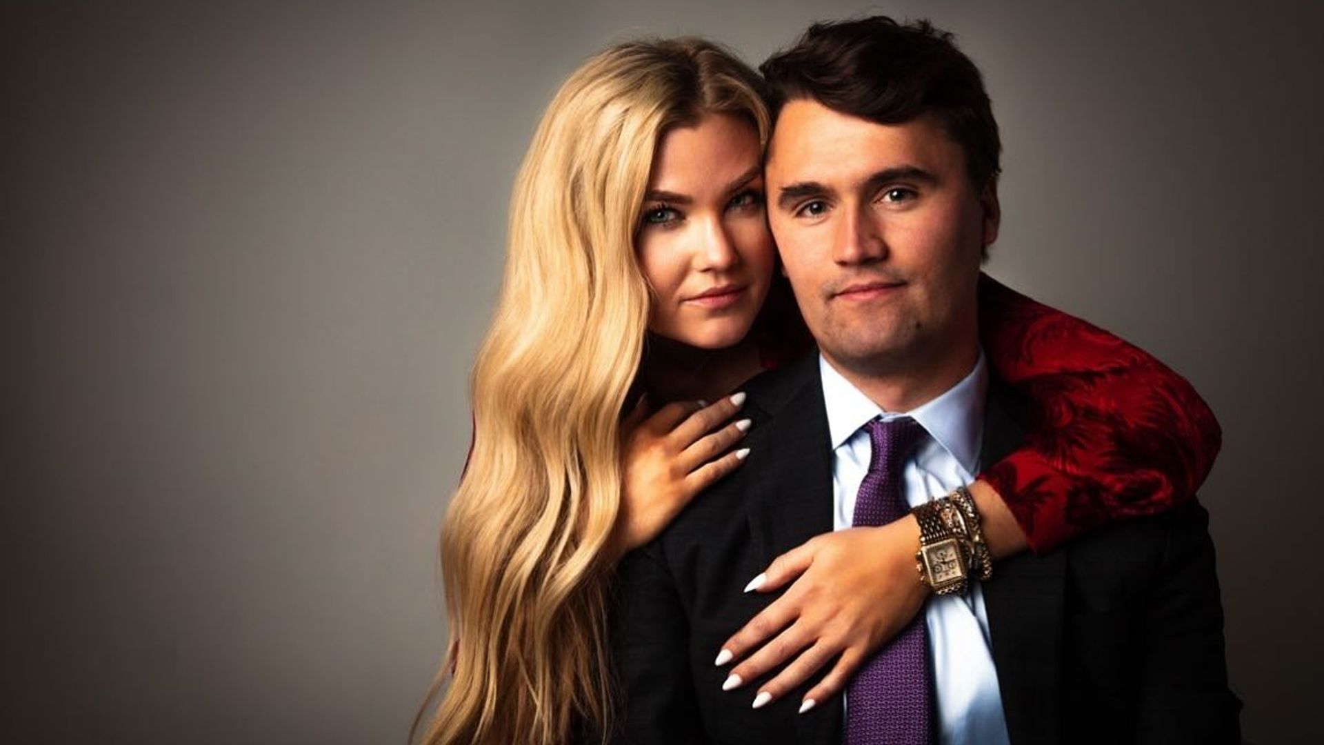 Charlie Kirk and Erica Frantzve embrace in front of the camera against a dark background