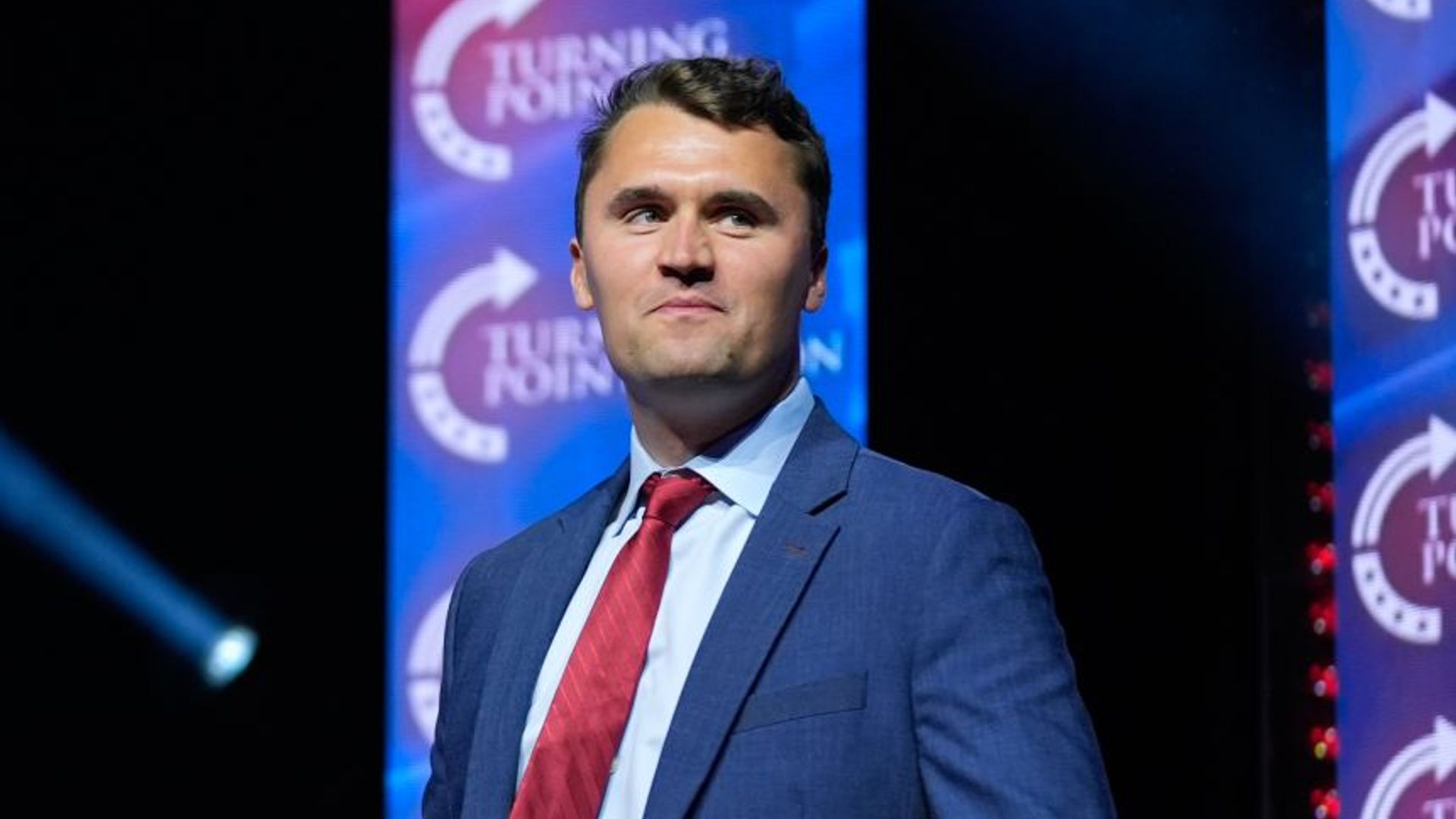 Charlie Kirk proudly stands against a dark background wearing a blue suit