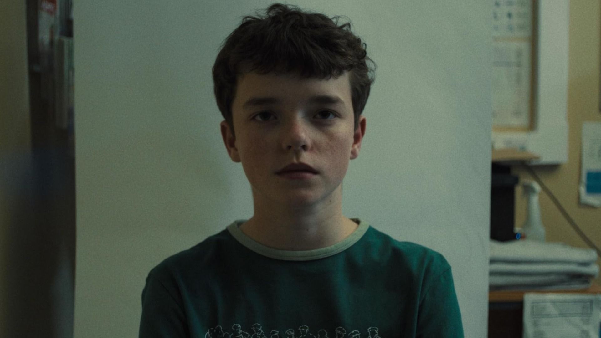 A boy with dark hair wearing a green t-shirt stands against a white wall