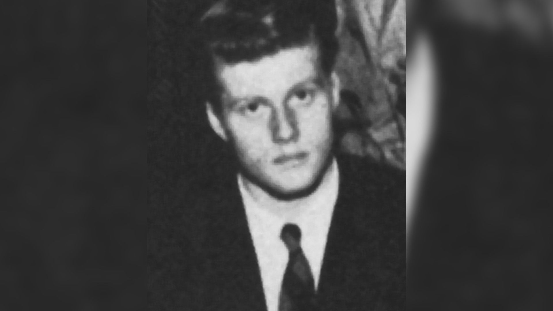 A black-and-white portrait of young Robert Redford in a suit and tie, looking directly at the camera.