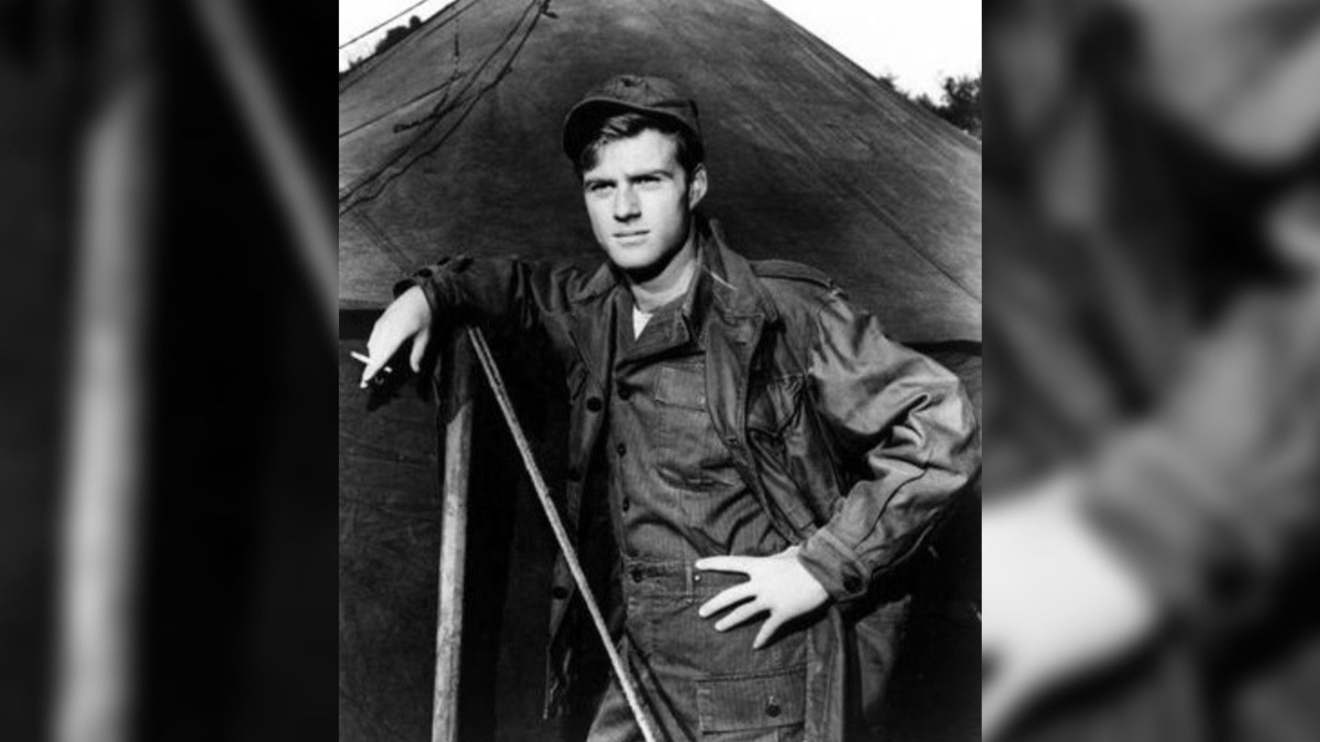Black and white photo of young Robert Redford in military uniform standing by a tent, scene from the film "War Hunt"