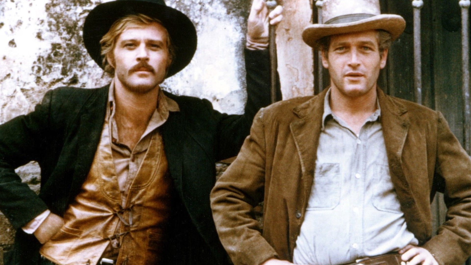 Robert Redford and Paul Newman in cowboy hats and jackets standing side by side against a wall, posing for a photo