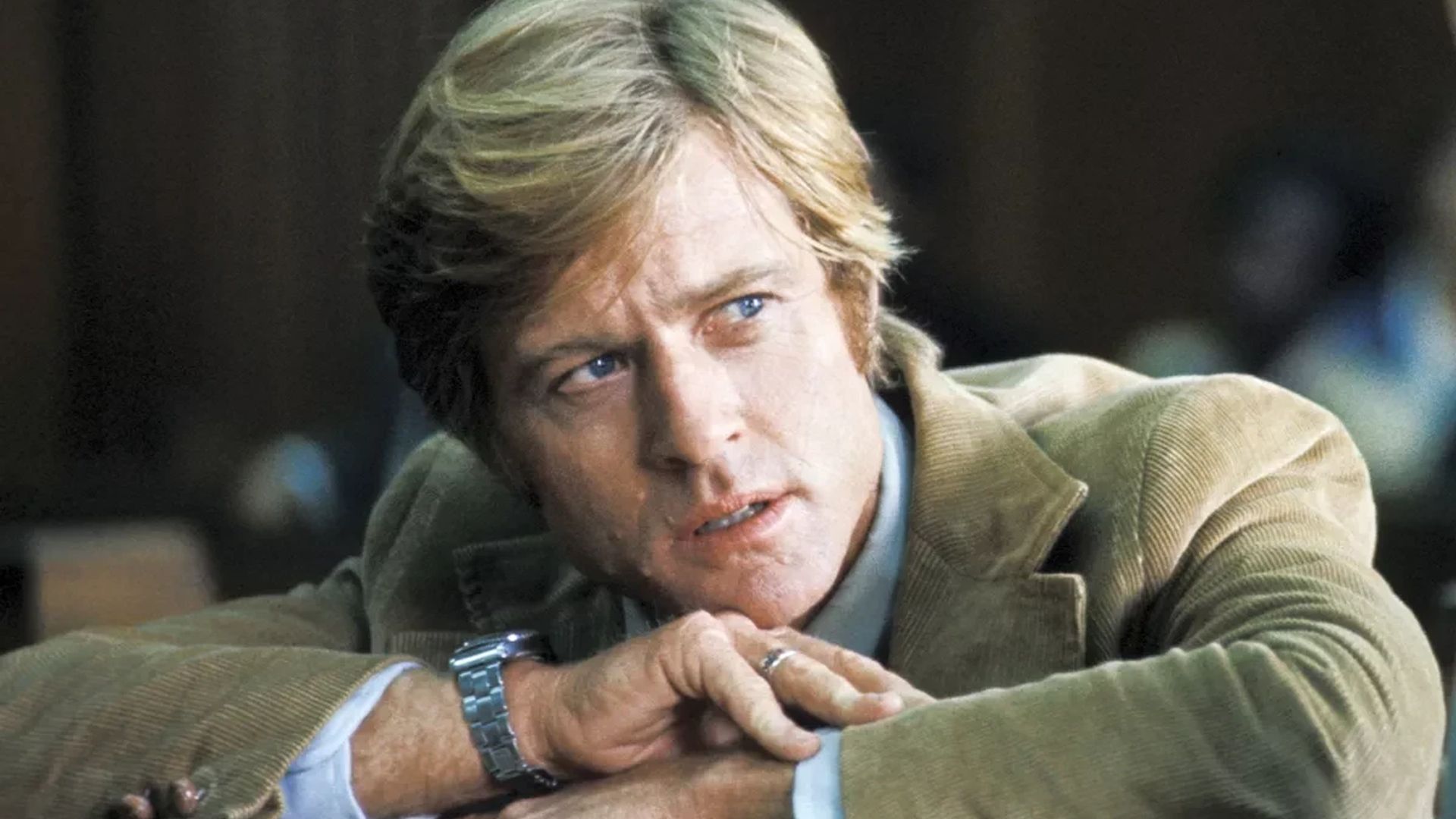 Robert Redford in a brown jacket with a watch on his wrist leans on his hands and looks to the side