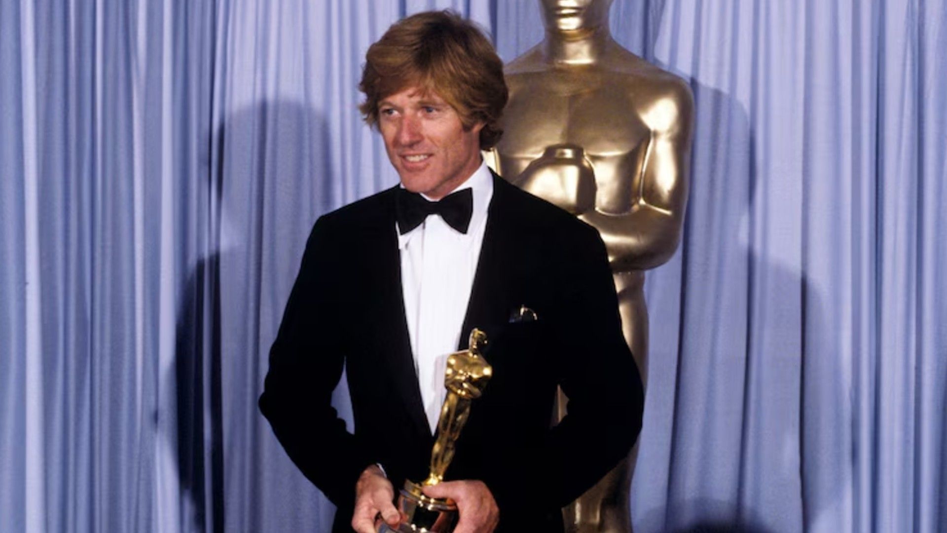 Robert Redford in a suit stands on stage holding an Oscar