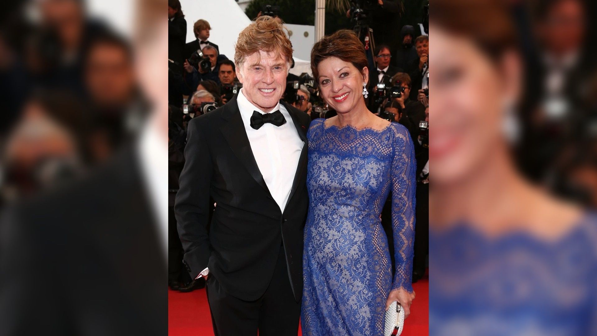 Robert Redford in a tuxedo and Sibylle Szaggars in a blue lace dress pose on the red carpet surrounded by photographers