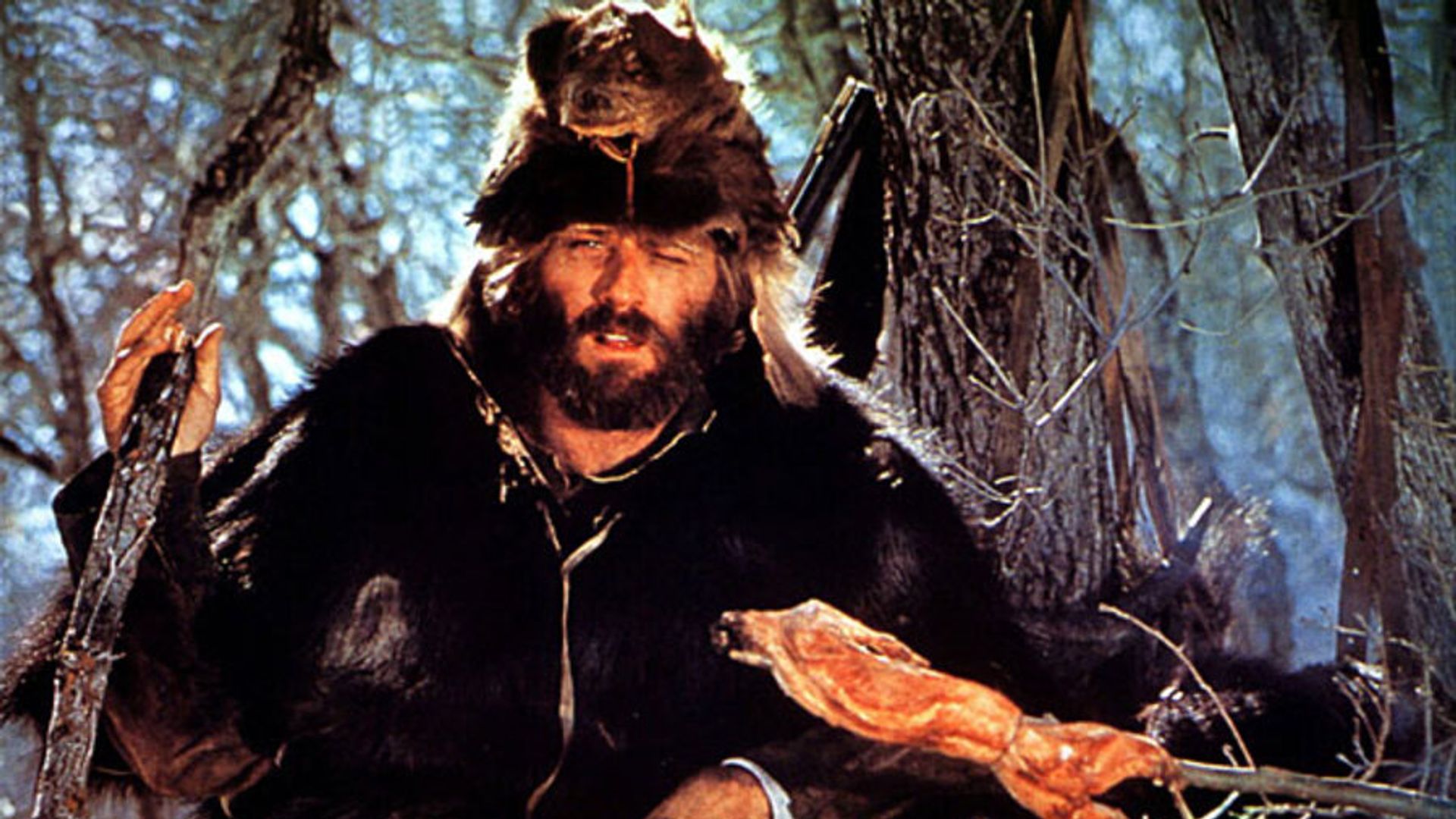 Robert Redford in fur clothing and a beast head cap grilling meat over a campfire in the forest, scene from the film "Jeremiah Johnson"