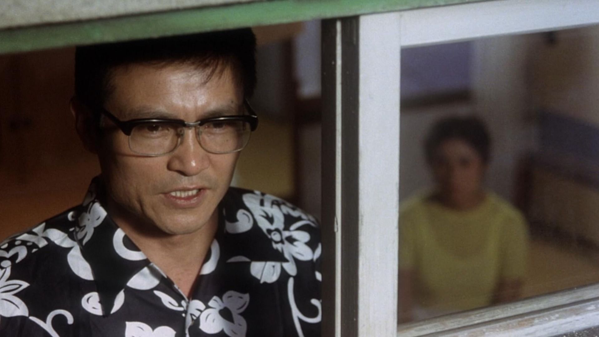 A man in glasses and a black shirt stands in front of a window with a girl sitting behind him, she's blurred
