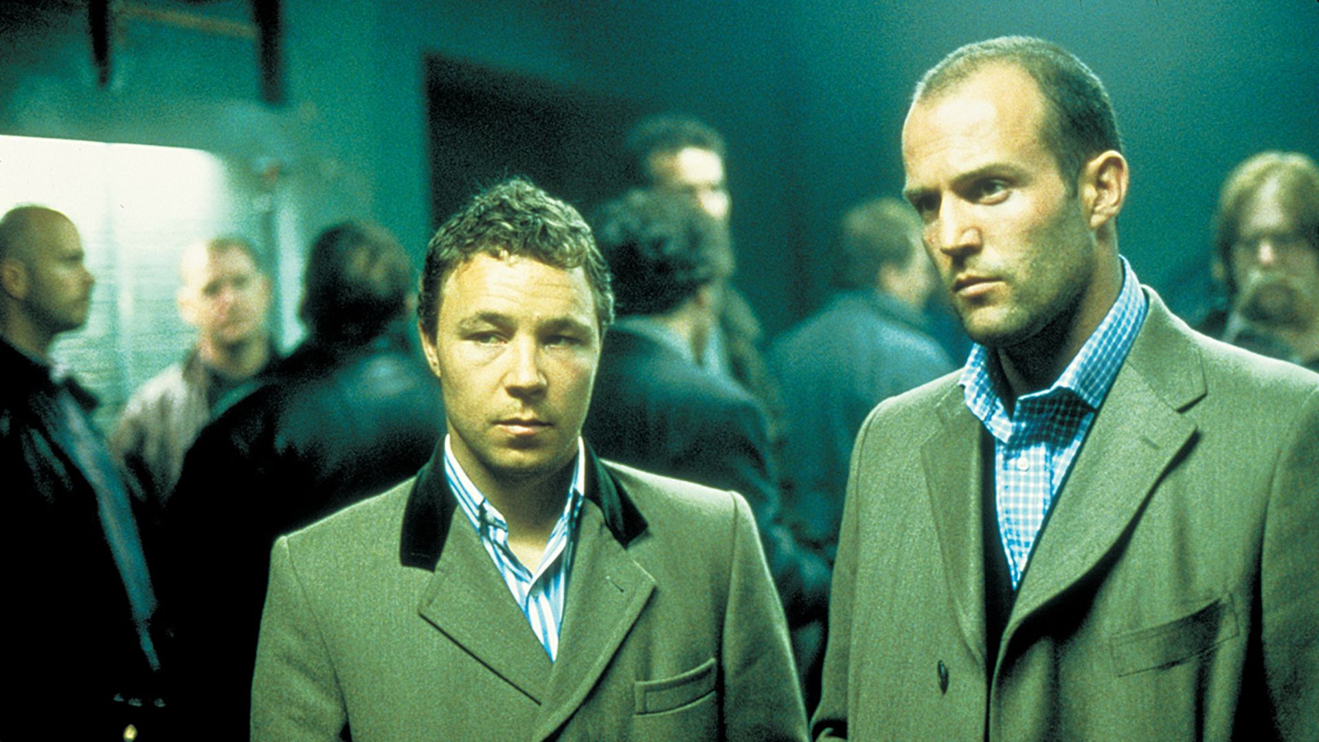 Steven Graham and Jason Statham, wearing coats, stand among men