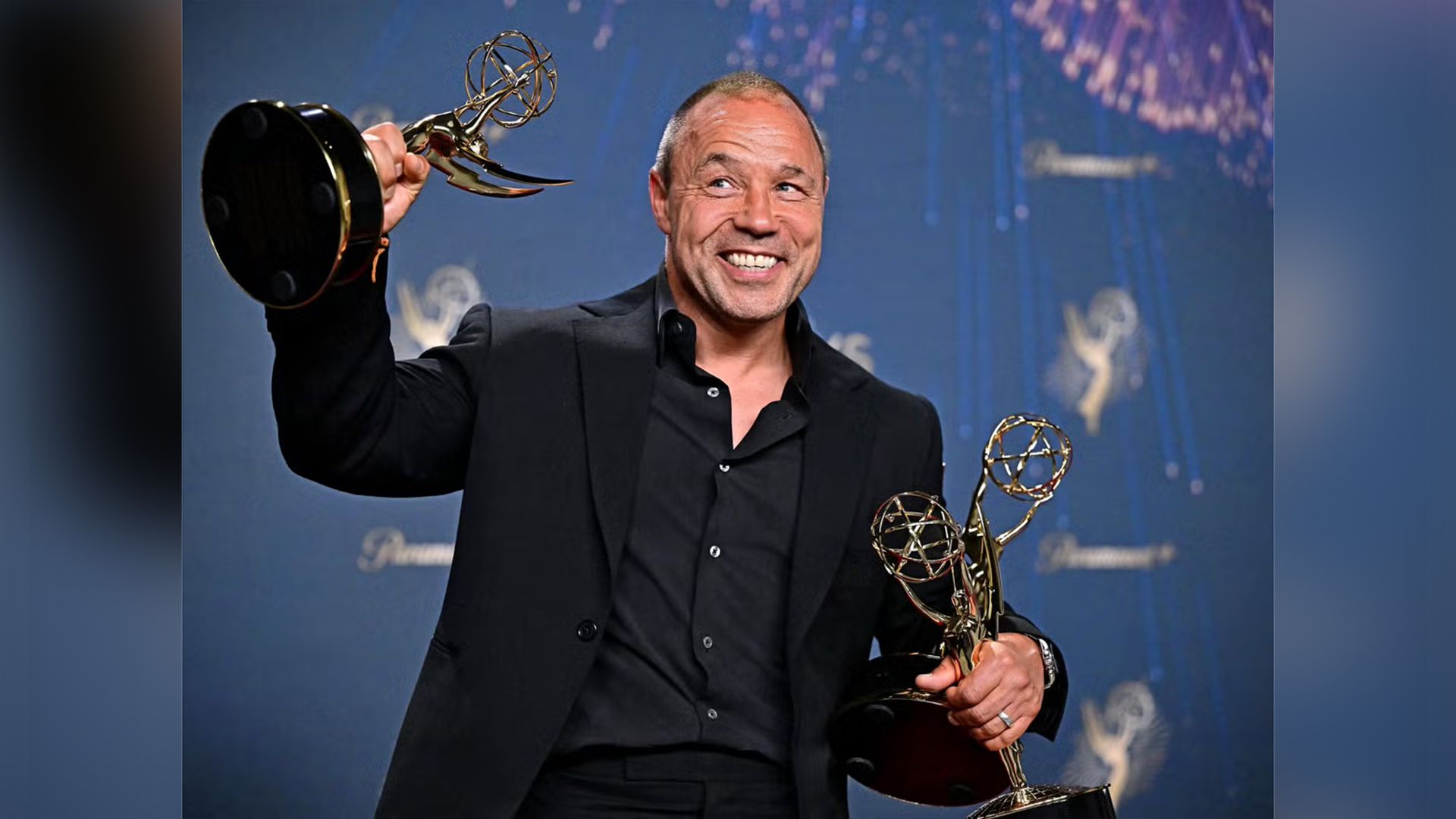 Stephen Graham in a black suit holds three golden Emmy statuettes in his hands, smiling widely