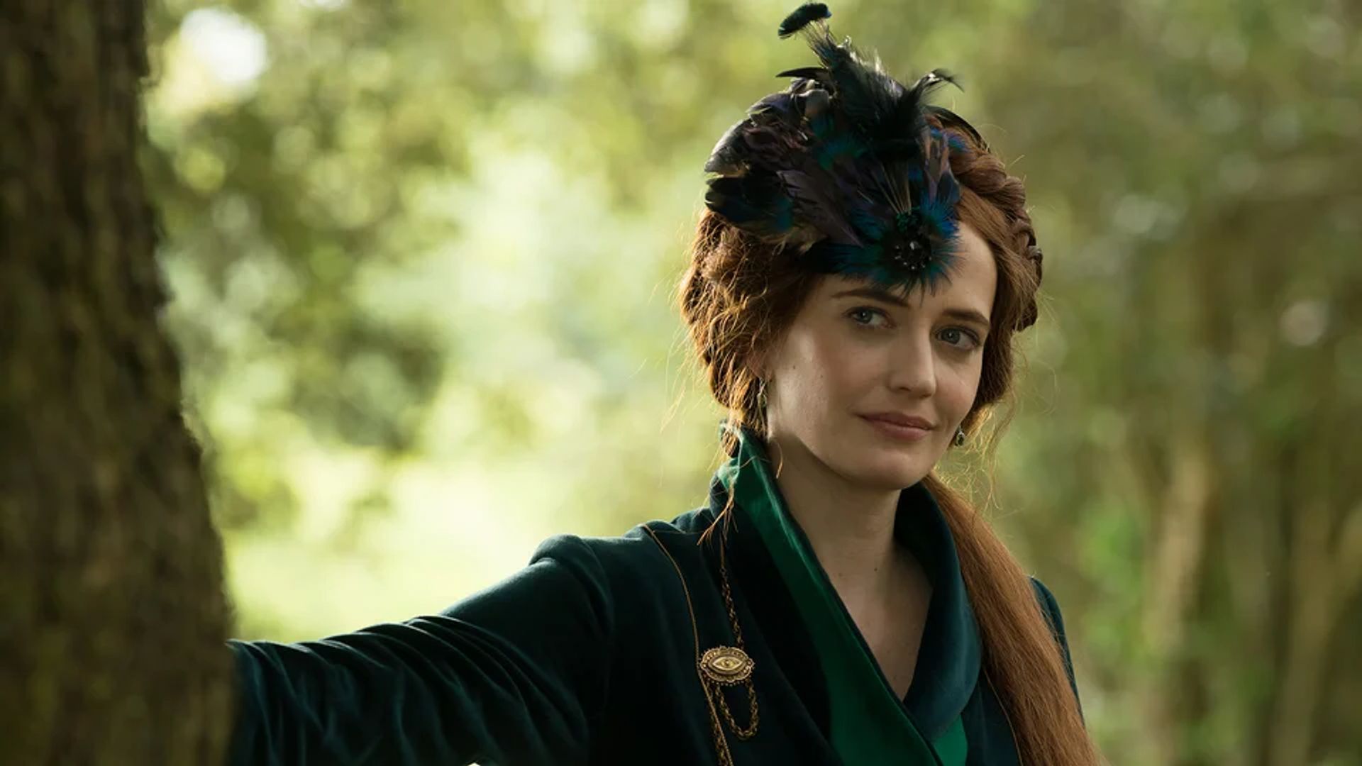 Eva Green in a dark green outfit with feather ornaments on her head stands by a tree against a forest backdrop