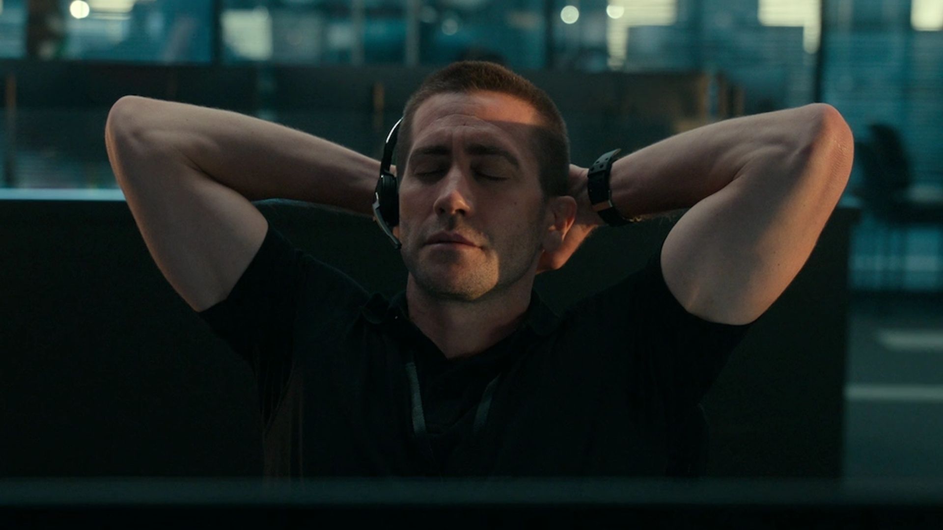 Jake Gyllenhaal wearing headphones with microphone sits with eyes closed, hands behind his head