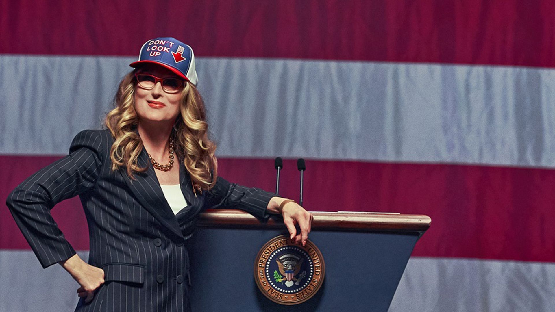 Meryl Streep in a suit and cap with American flag stands against a backdrop of the US flag, leaning on a podium