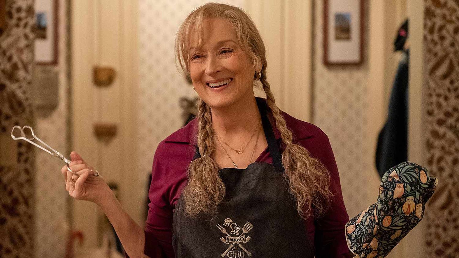 Meryl Streep with braids holds tongs in one hand, wearing a kitchen mitt on the other