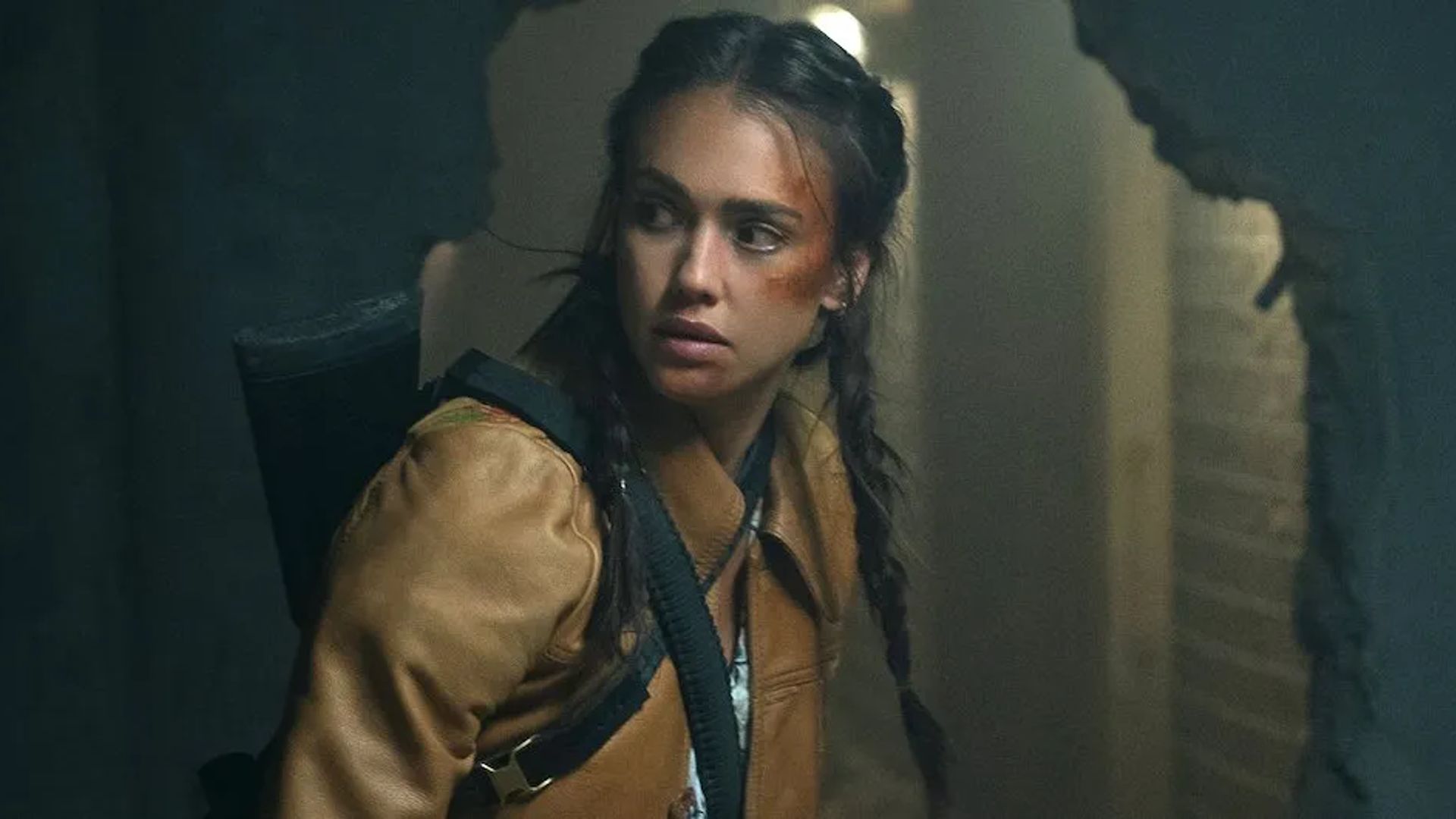 Jessica Alba with braided hair and a backpack on her shoulder looks warily to the side, standing by a broken wall