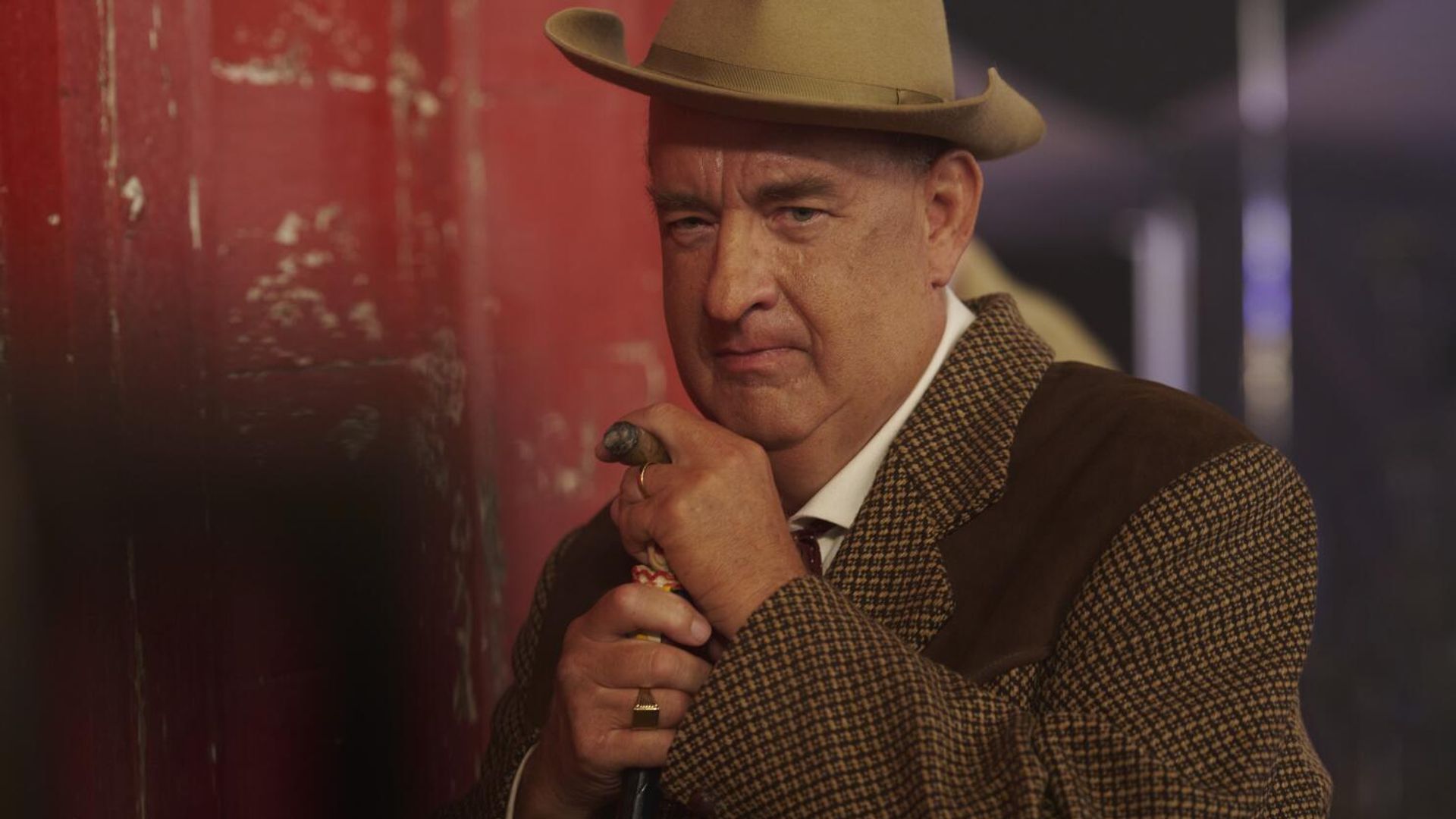 Tom Hanks in brown suit and hat leans on a cane, looking seriously into the camera