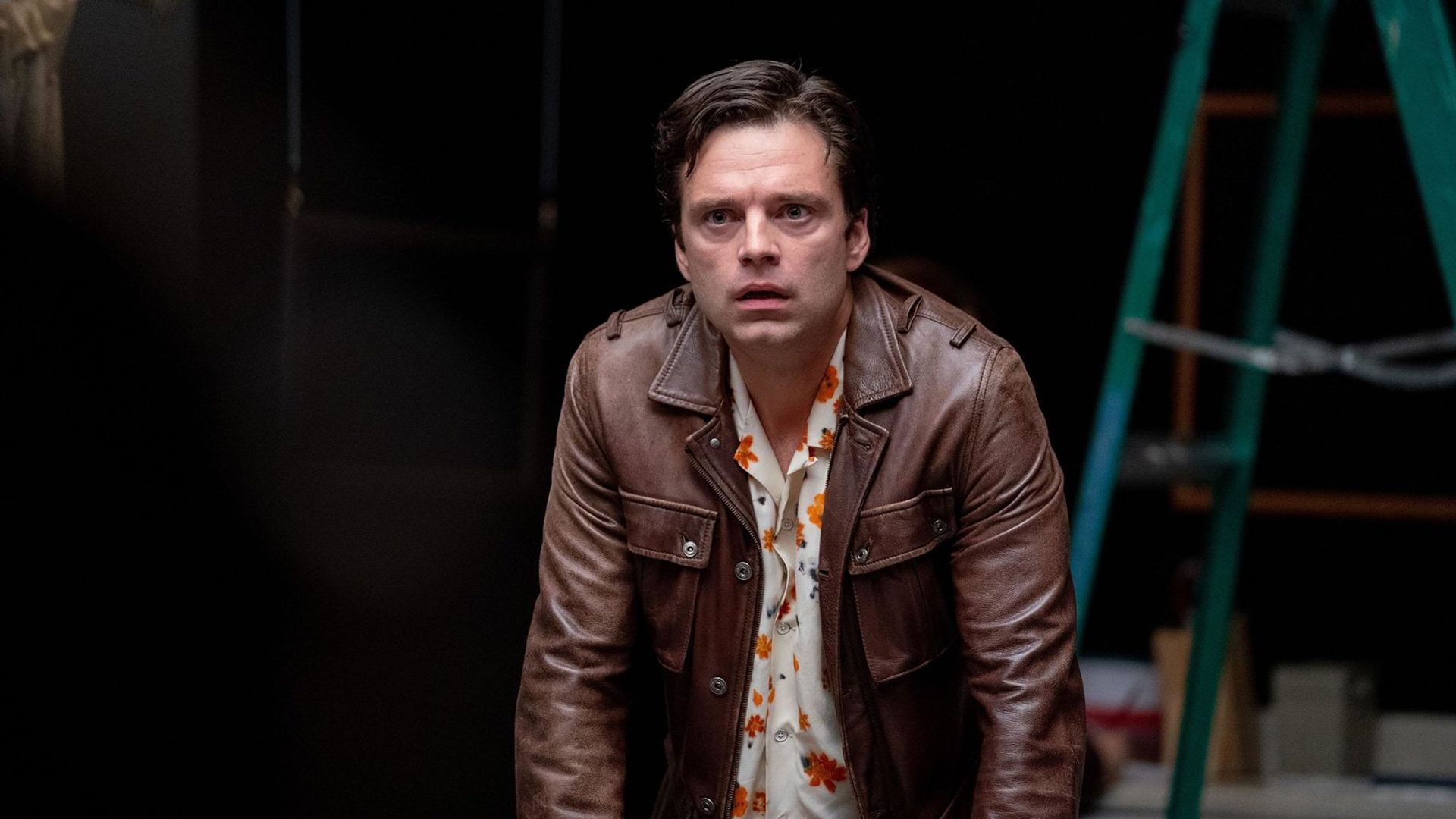 Sebastian Stan in a brown jacket looking ahead against a black background
