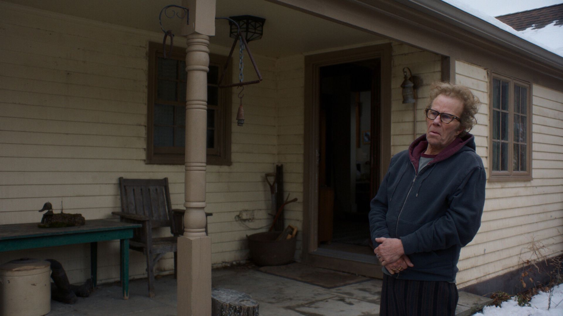 Tom Waits in a blue hoodie stands near a wooden house