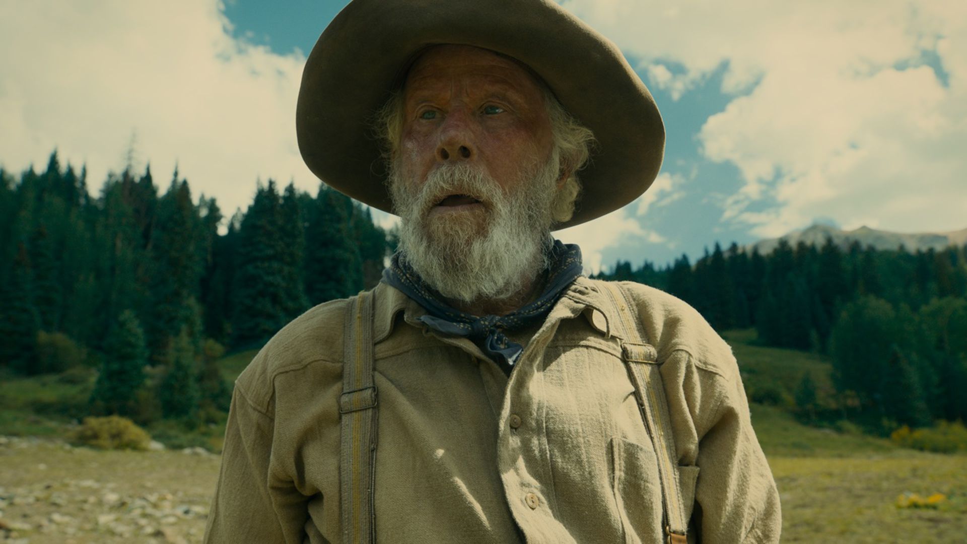 Tom Waits in a hat, green shirt and blue neckerchief against a green valley backdrop