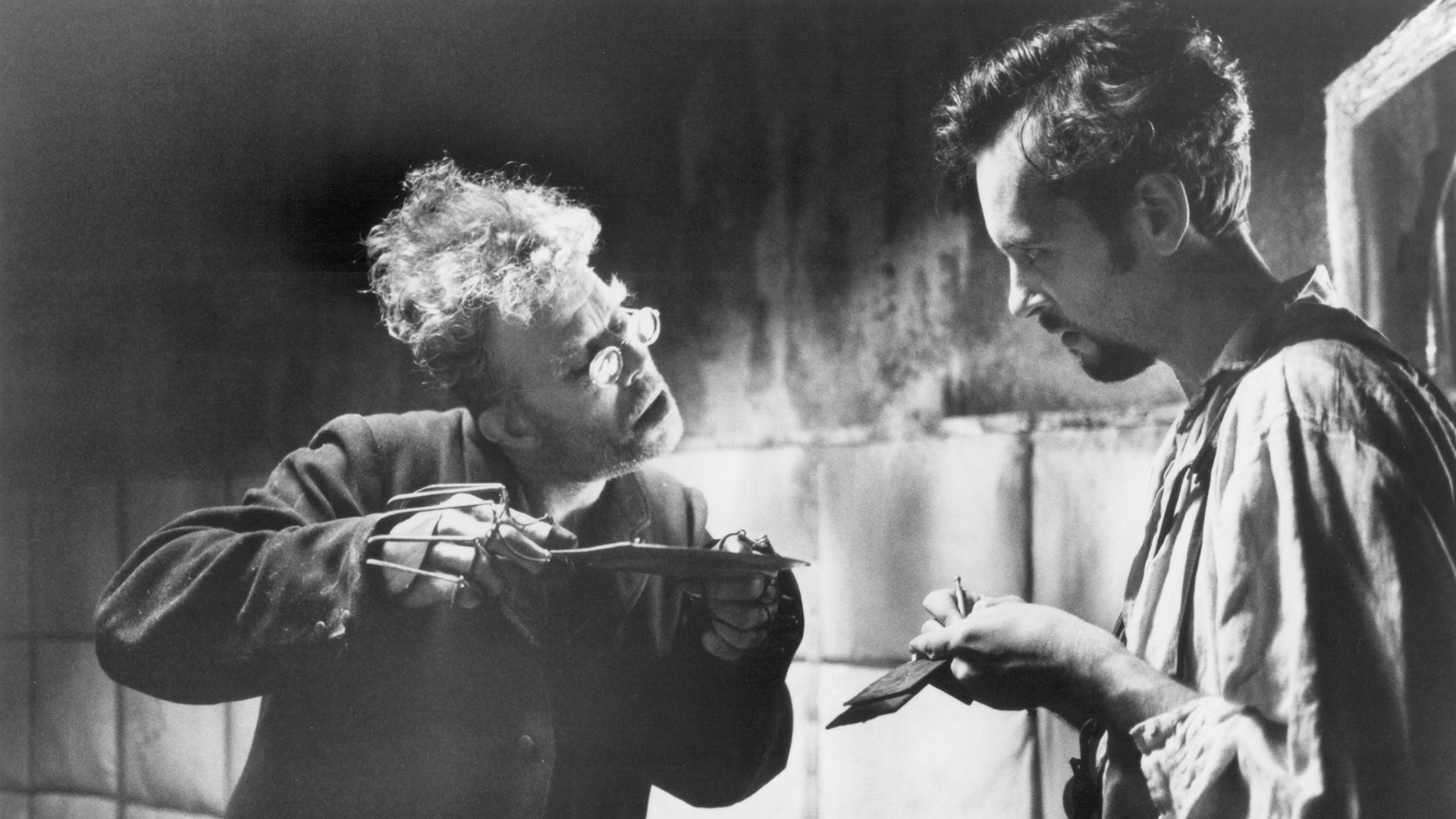 Tom Waits in round glasses and with finger braces holds a saucer in his hands, next to him stands a young man with a notepad