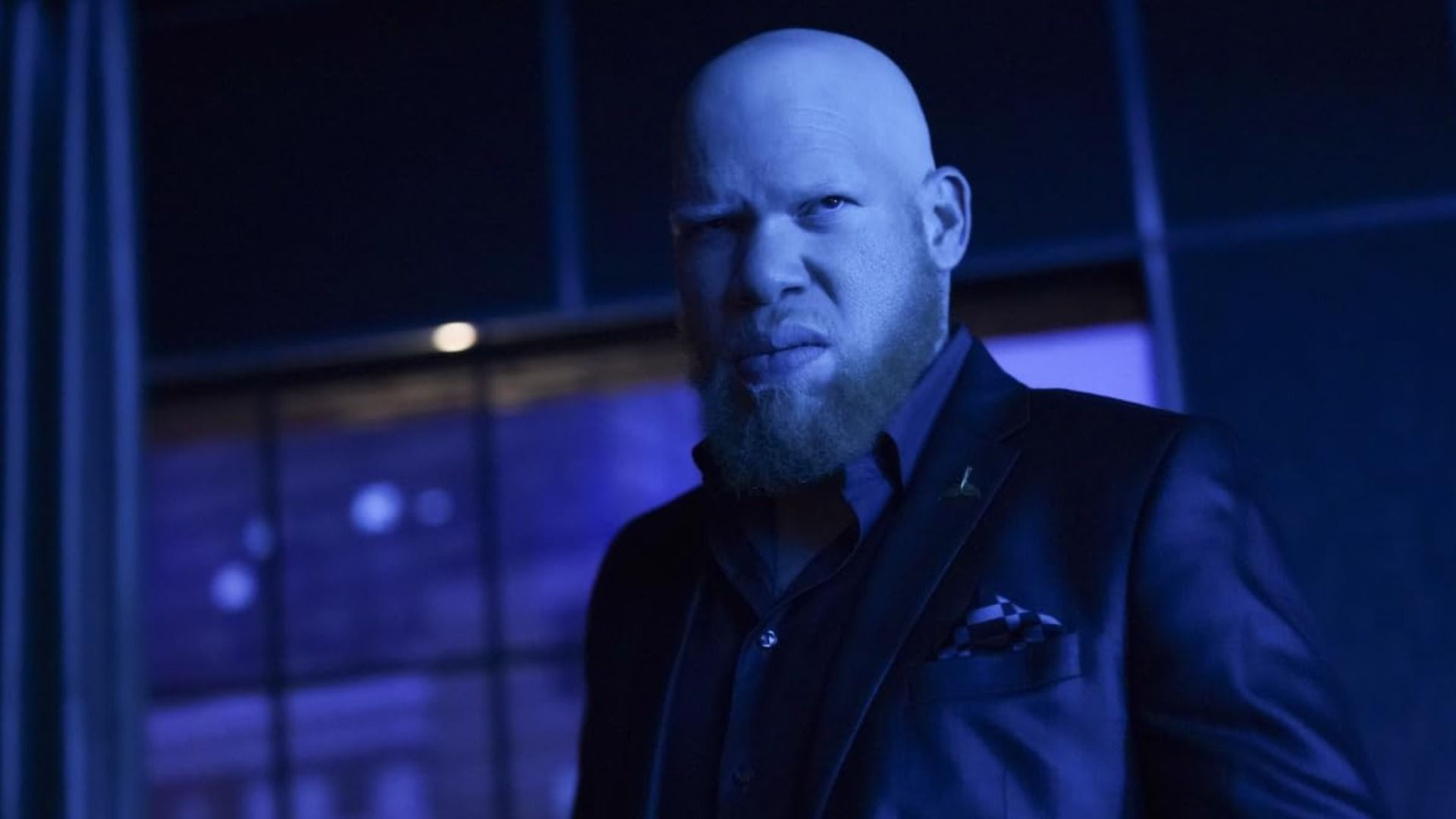 A bald man in a black suit stares menacingly to the side