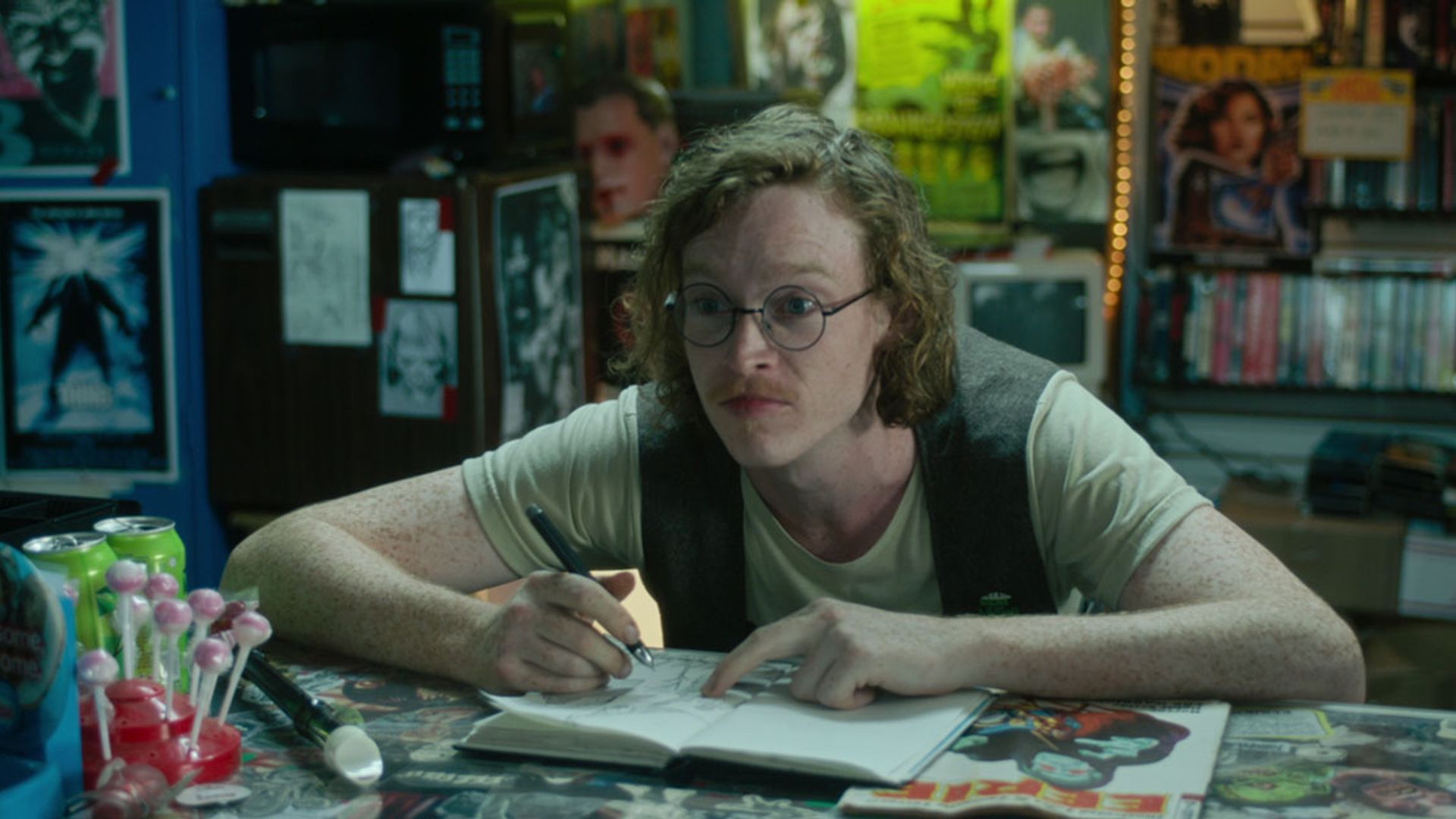 Caleb Landry Jones wearing glasses and a vest sits at a table with a notepad, drawing and looking to the side