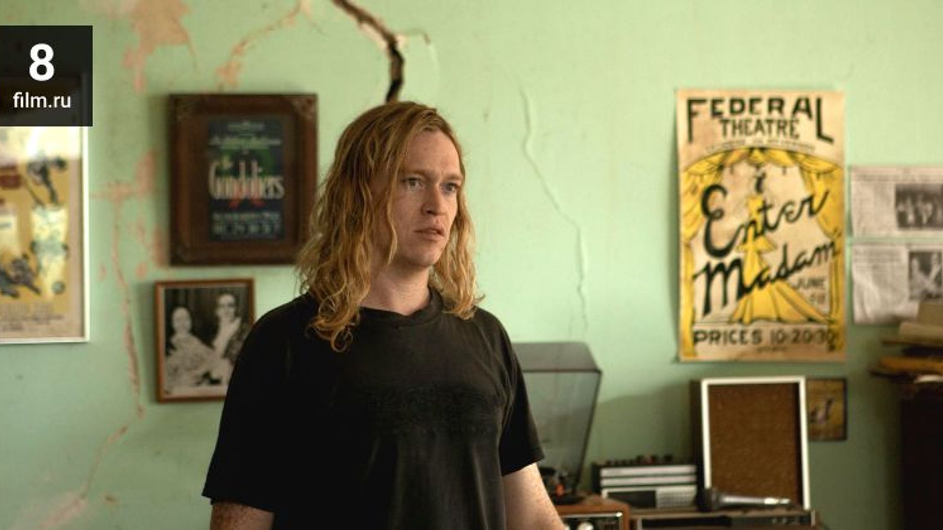 Caleb Landry Jones with long blonde hair in a black t-shirt stands in a room with posters on the wall