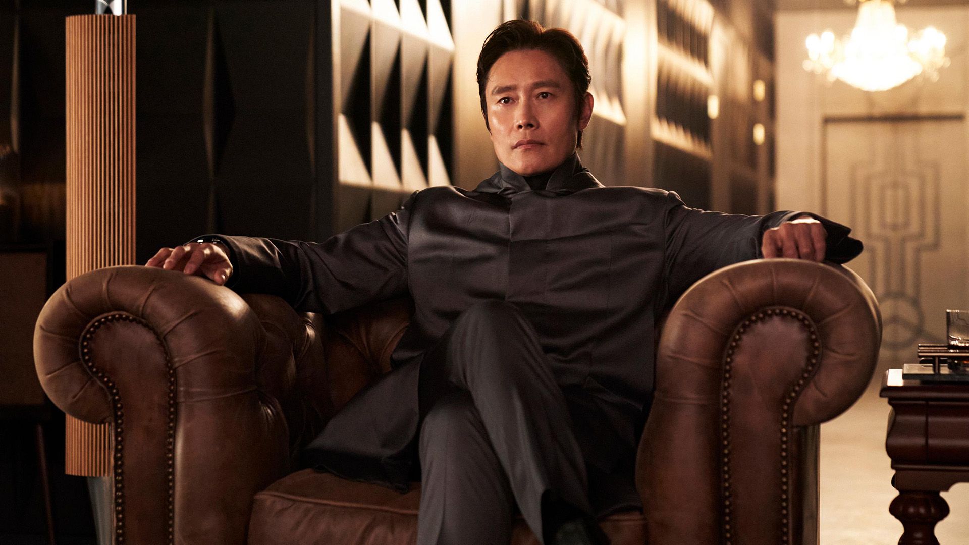 Lee Byung-hun in a gray blazer sits in a leather armchair with his legs crossed