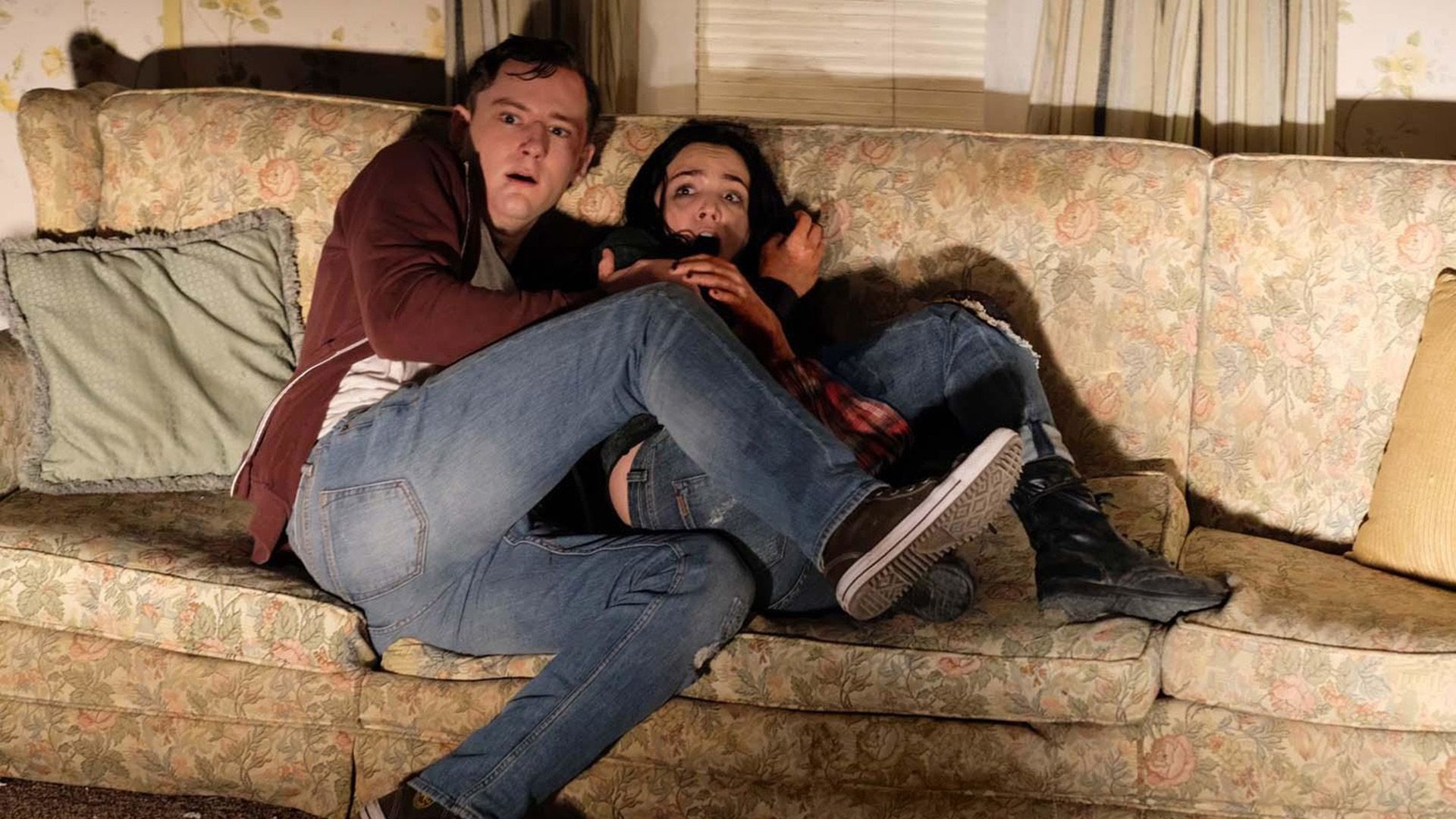 Lewis Pullman and a girl sitting terrified on a couch with their legs tucked up