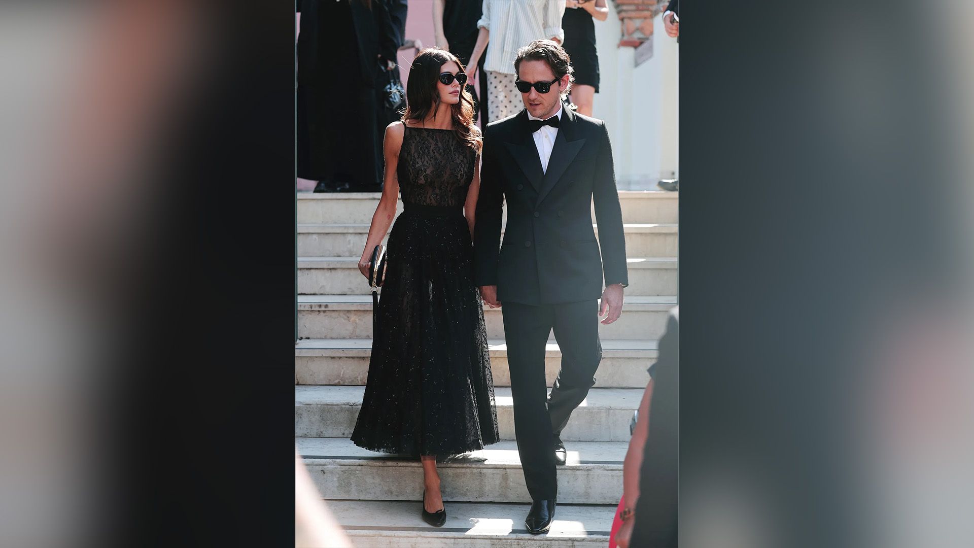 Lewis Pullman in a black tuxedo and Kaia Gerber in a black evening gown descend the staircase, both wearing black sunglasses