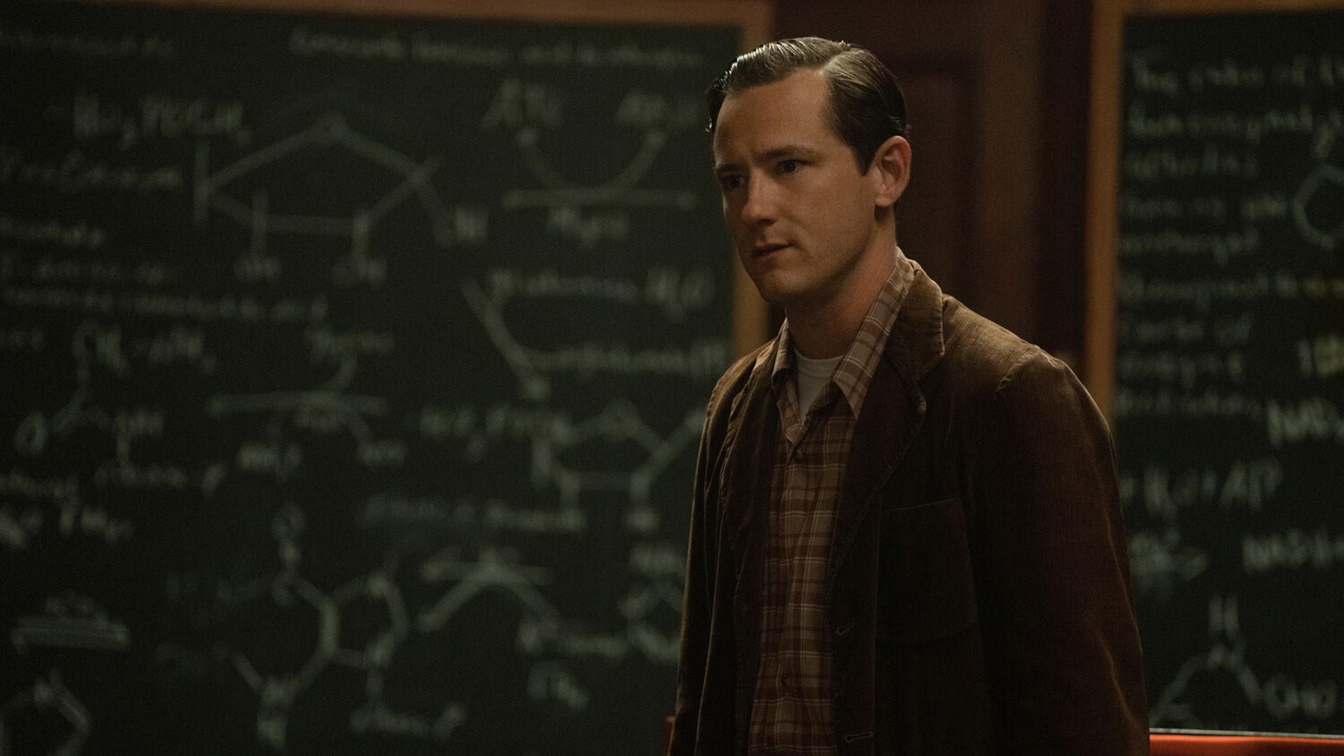 Lewis Pullman in a brown corduroy blazer and checkered shirt stands against a blackboard with chemical formulas