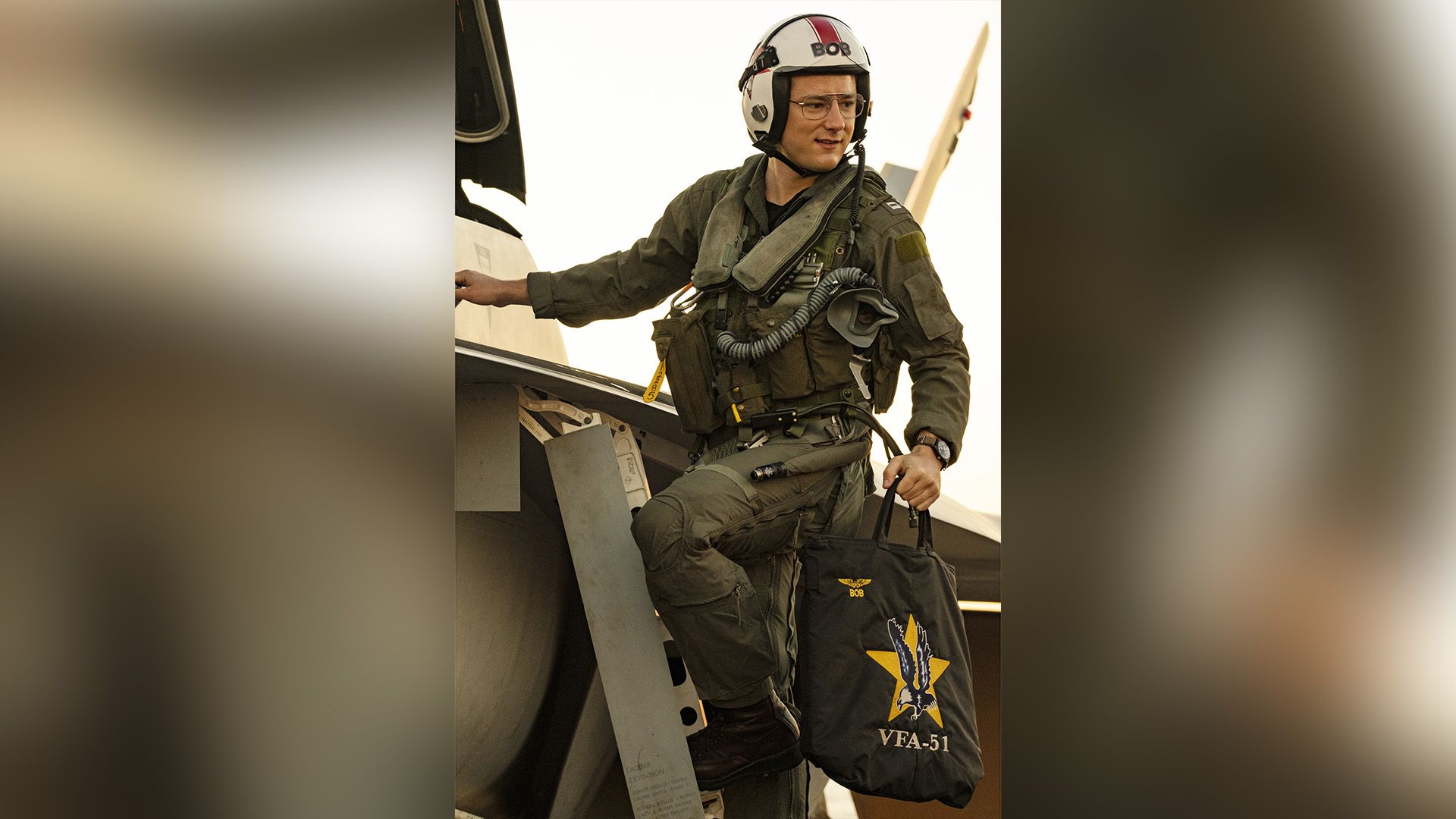 Lewis Pullman in military pilot uniform stands on the step to a fighter jet
