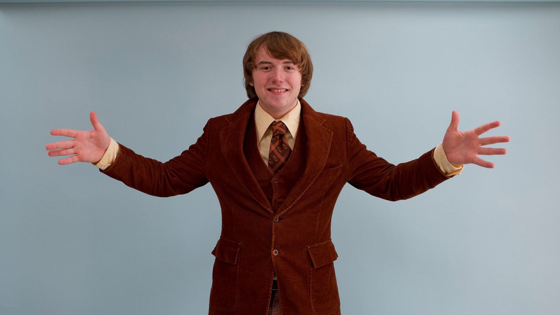 Young Cooper Hoffman in a brown corduroy jacket stands with his arms spread wide