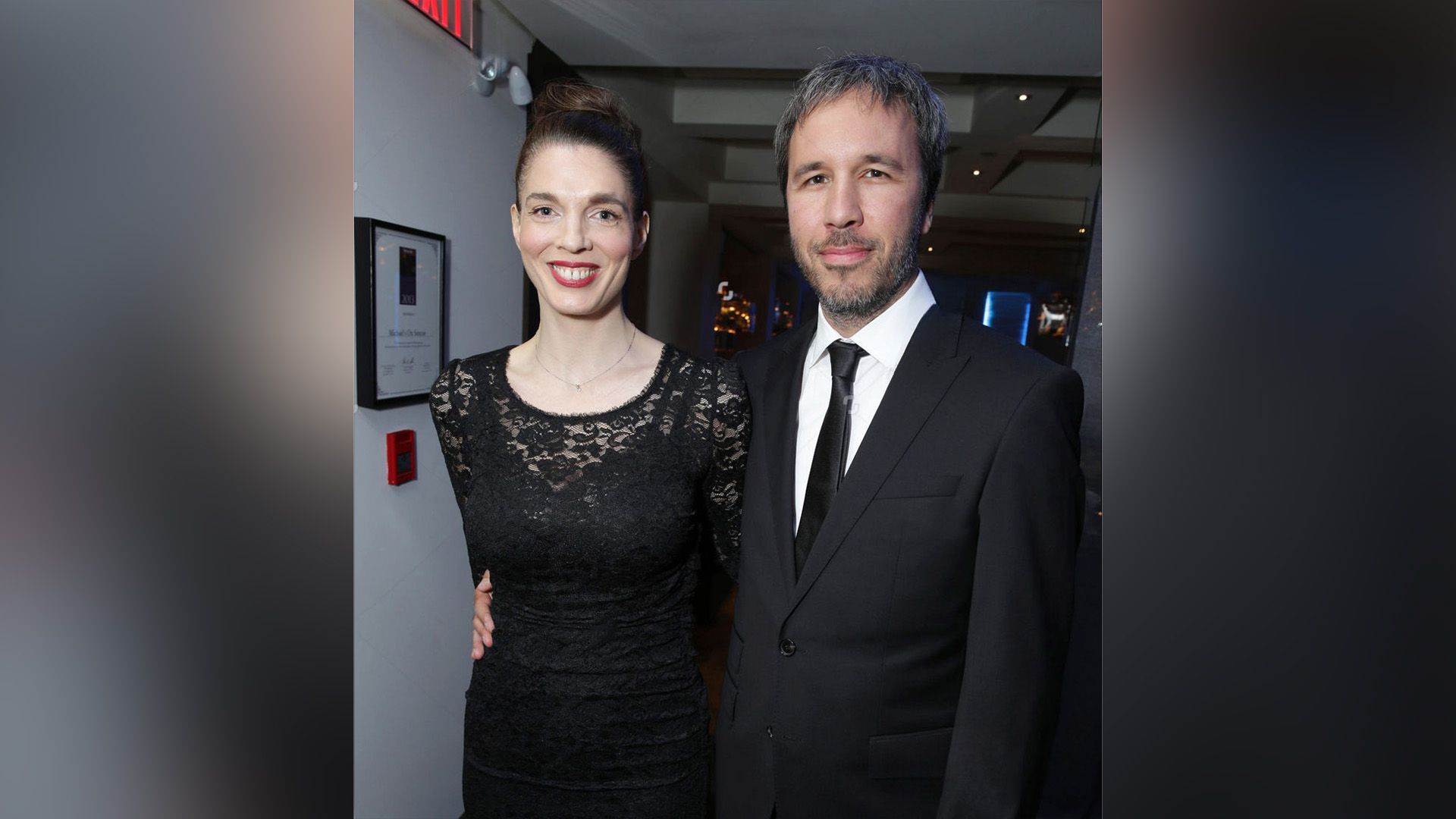 Denis Villeneuve in a suit embraces a smiling Masha Grenon in a black lace dress around the waist