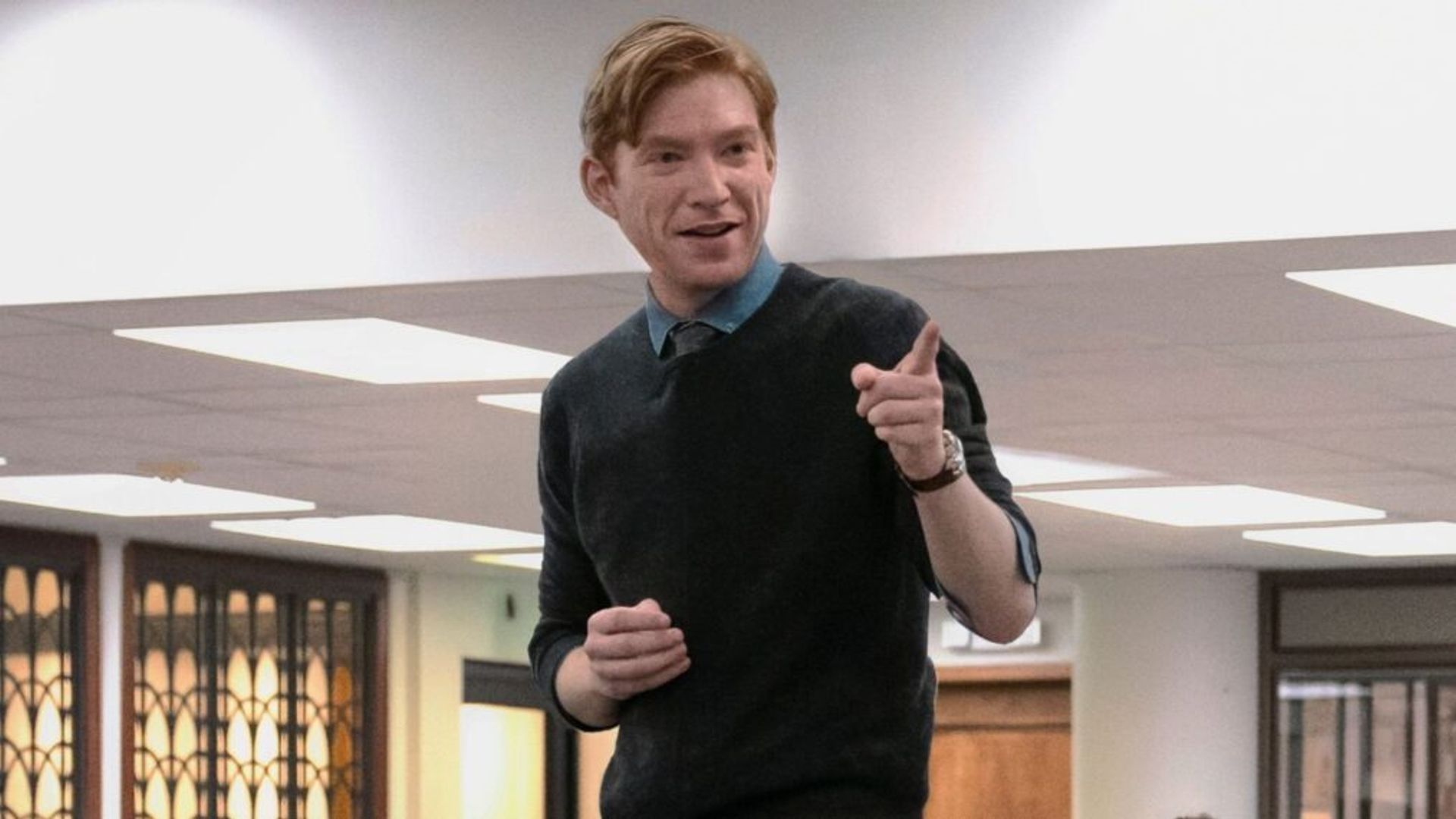 Domhnall Gleeson in a dark blue sweatshirt standing on a table and pointing his finger