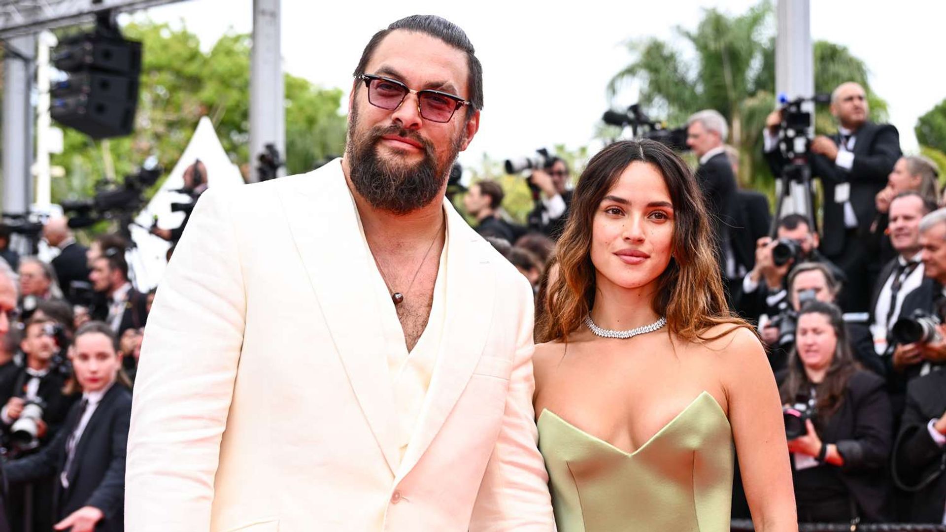 Jason Momoa in a white suit and Adria Arjona in an olive dress on the red carpet