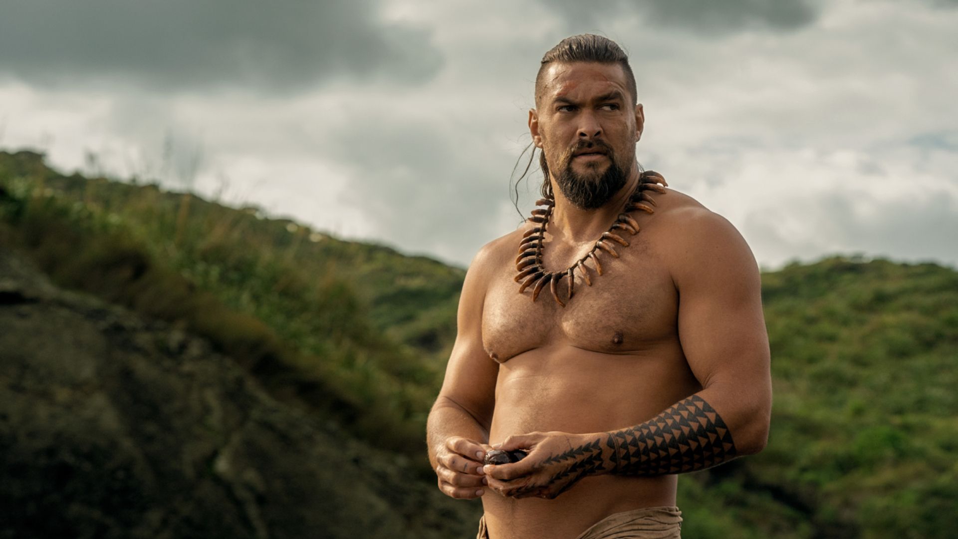 Jason Momoa shirtless with traditional Hawaiian decorations and tattoos against a mountain backdrop