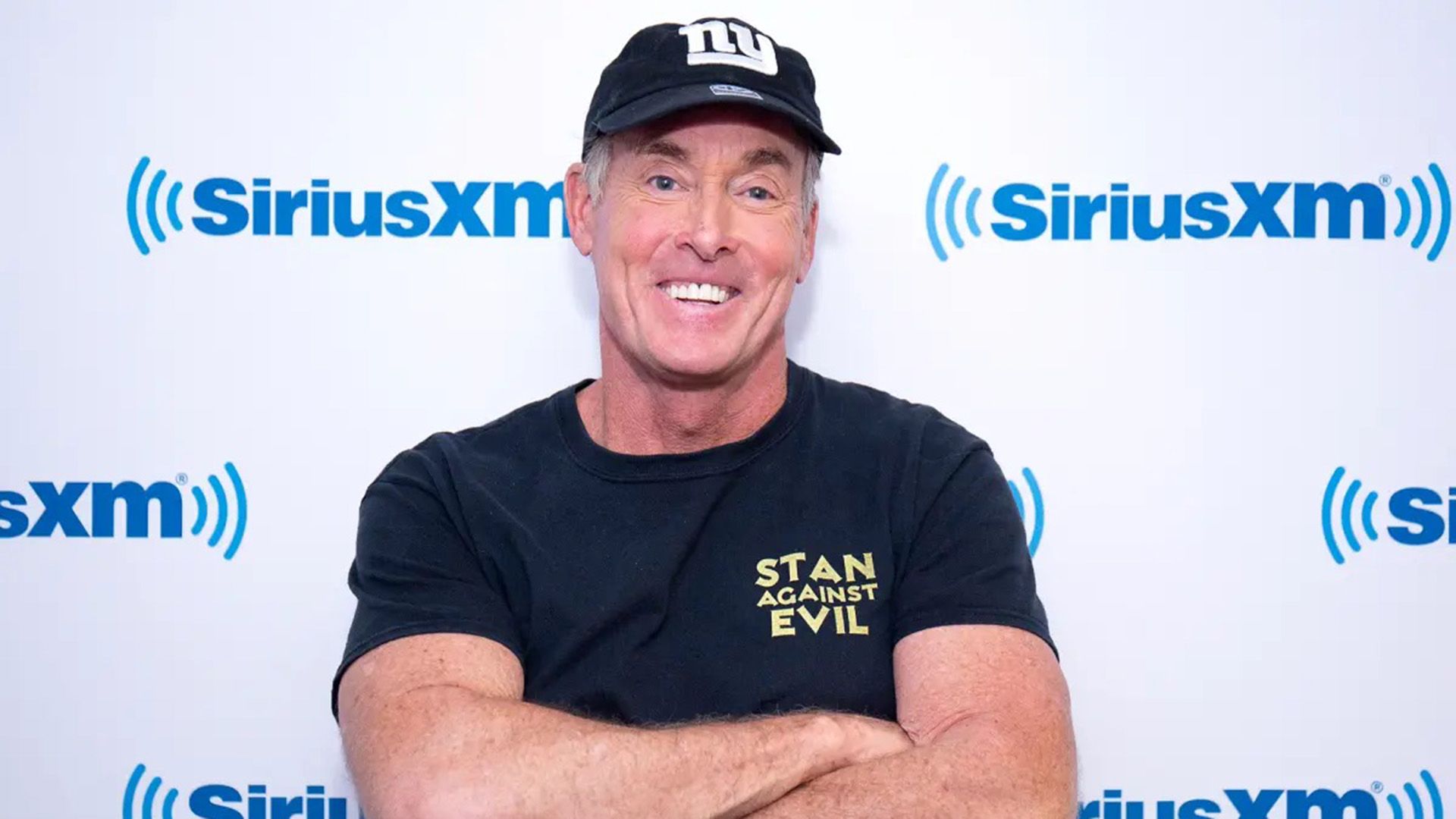 Actor John C. McGinley wearing a baseball cap, standing with arms crossed and grinning widely