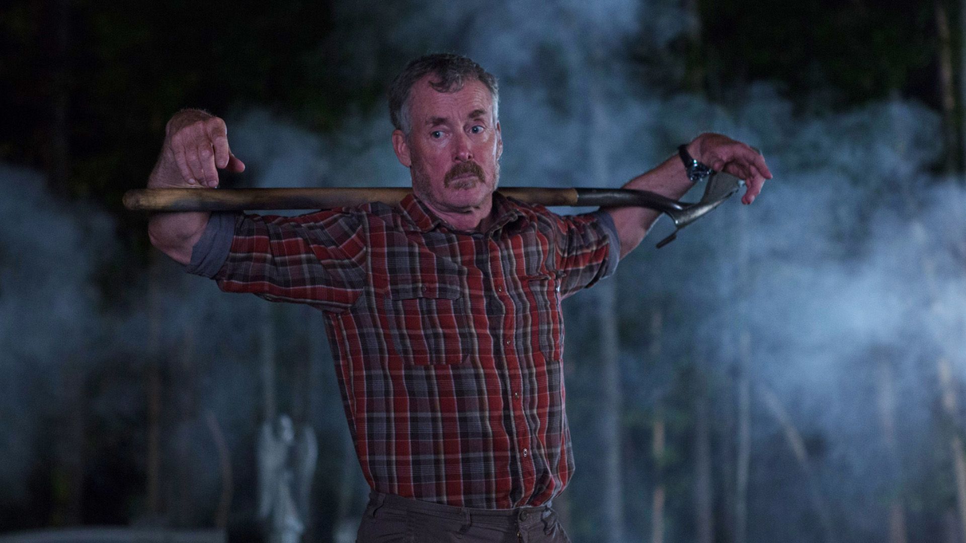 John C. McGinley in a red plaid shirt stands in the dim light with a shovel over his shoulders