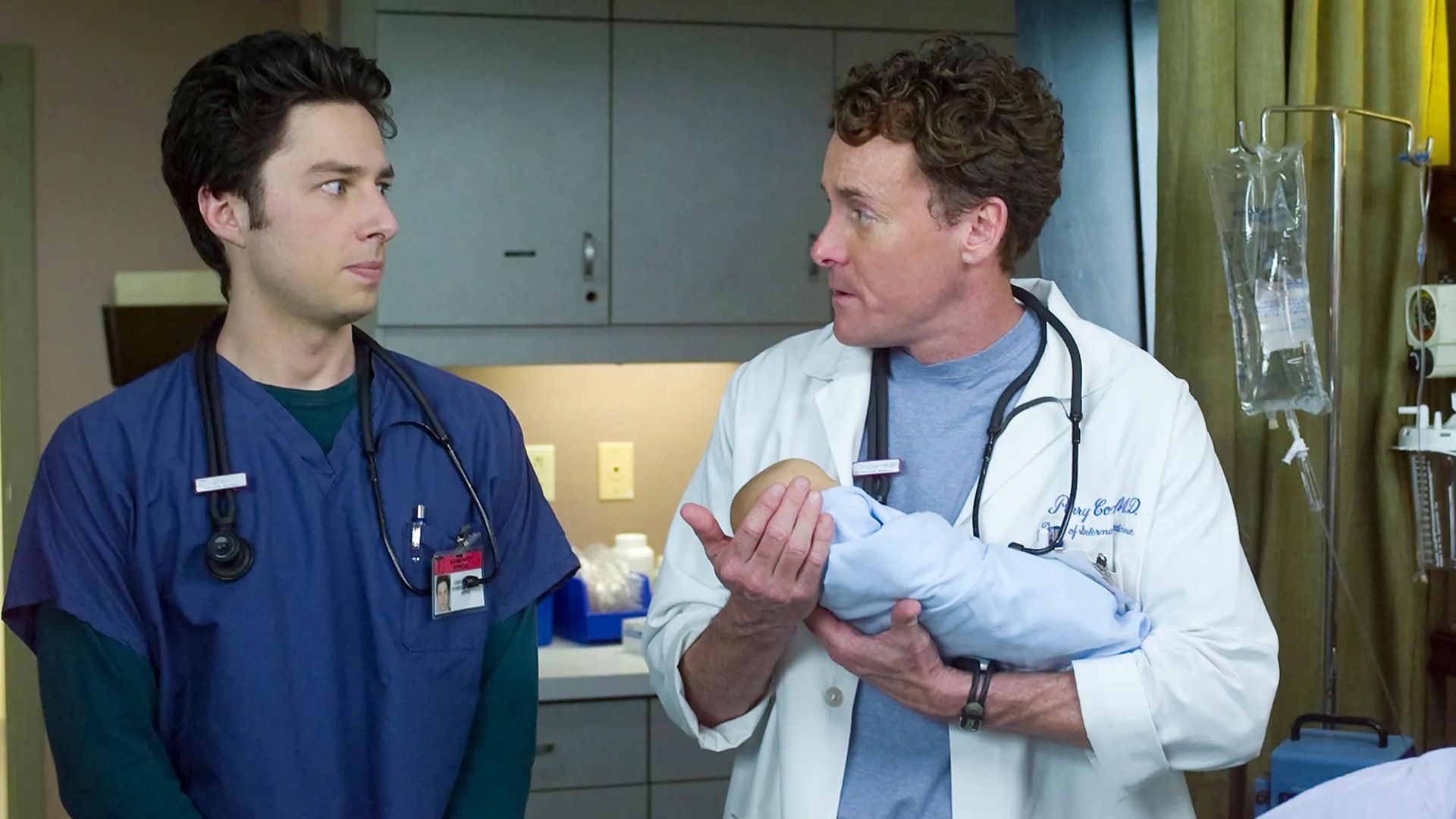 John C. McGinley in a white coat holding a baby, with Zach Braff standing nearby in a blue coat