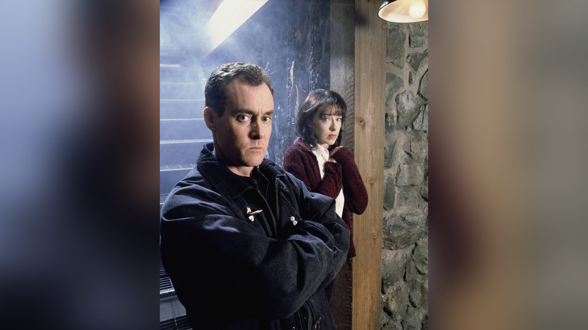 John C. McGinley with a serious expression stands near a staircase with a frightened girl behind him