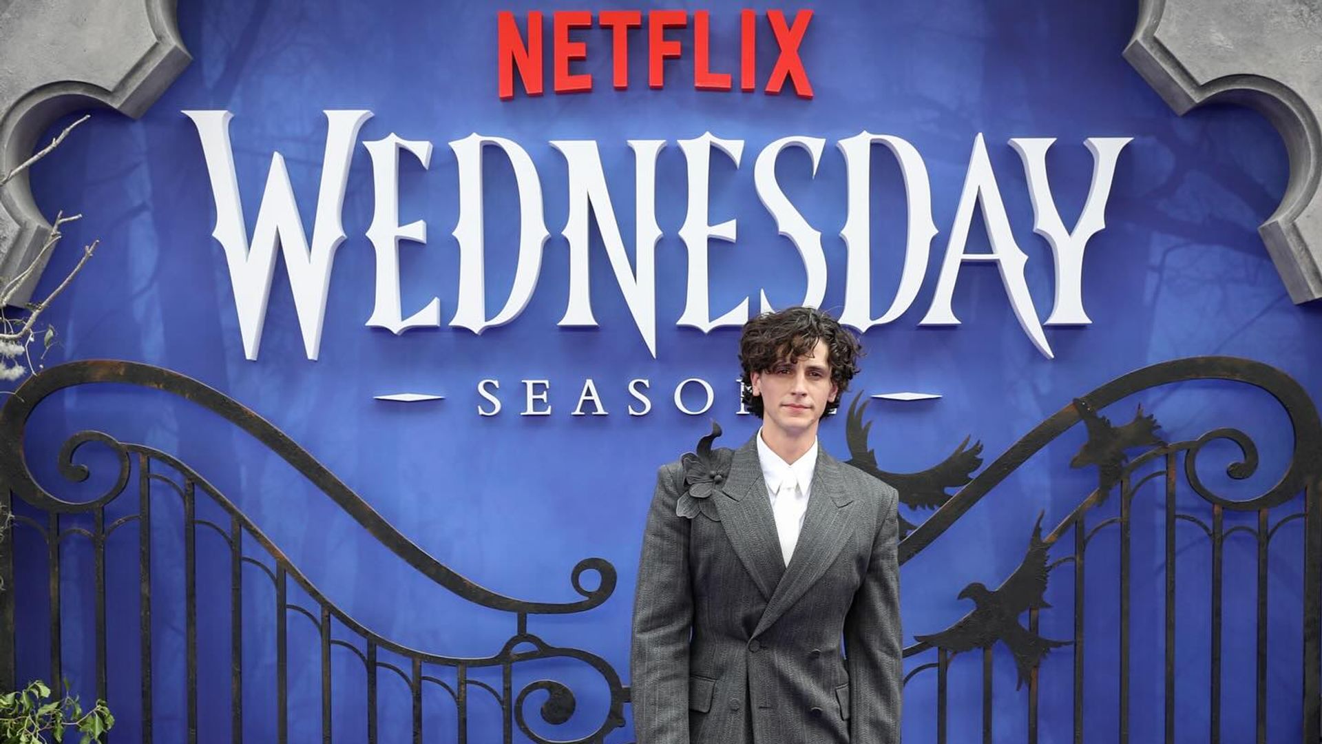 Owen Painter in a suit stands against a Wednesday Netflix photo wall backdrop